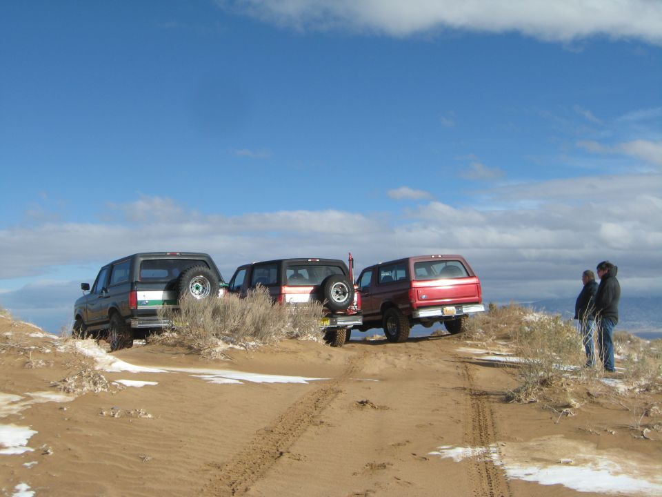 Three Ford Broncos parked on a sandy hill with two people standing nearby.