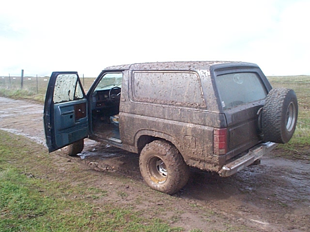 Mud-covered dark Ford Bronco SUV with driver's door open and rear-mounted spare tire on a dirt road