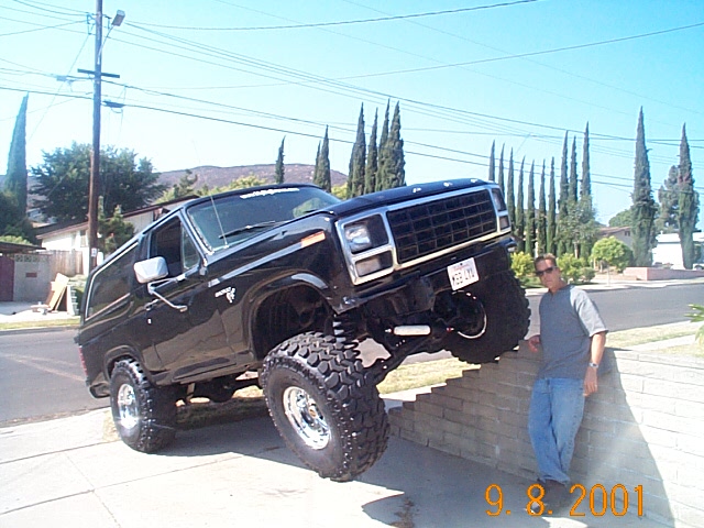 Black lifted full-size Ford Bronco with front wheels up on a brick wall, man standing beside it on a street