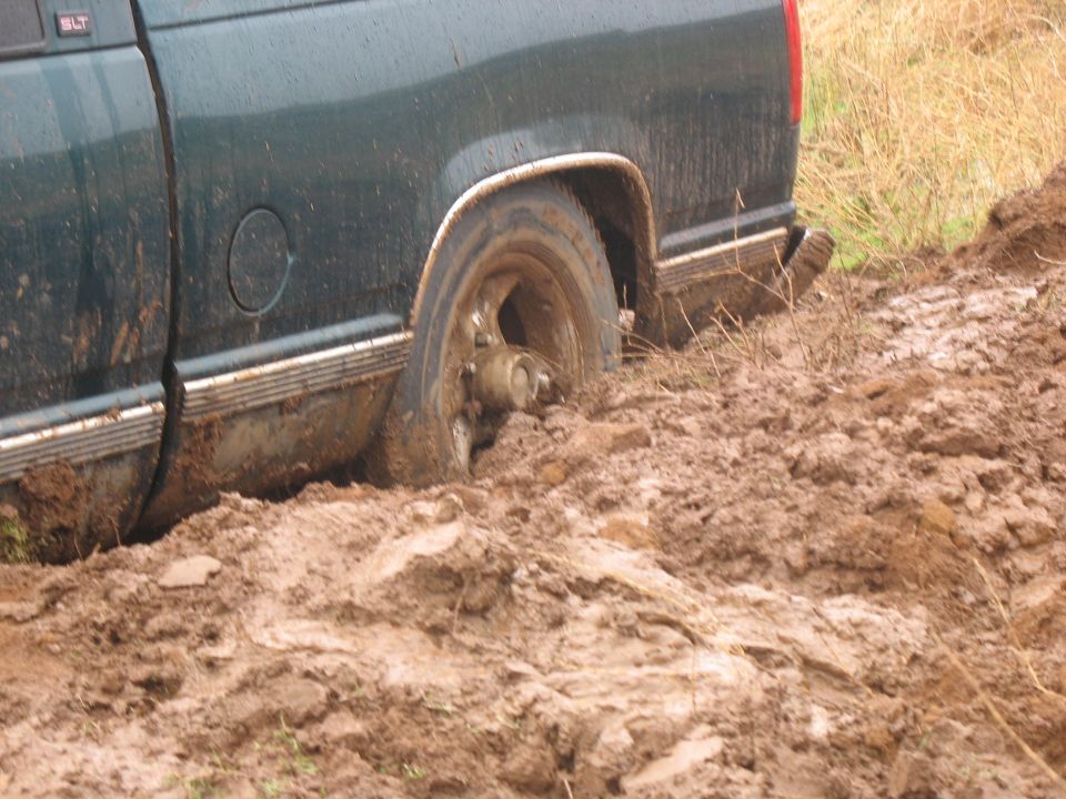 Rear driver's side wheel of a dark green pickup truck stuck in mud.