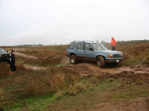 2001 Ford F-250 - A light blue SUV is driving through a muddy terrain with a person in an orange hoodie standing in the background. Another vehicle is partia…