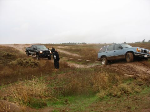 2001 Ford F-250 - A blue SUV is being pulled out of a muddy ditch by a black pickup truck on a cloudy day. Two people are standing near the pickup truck.
