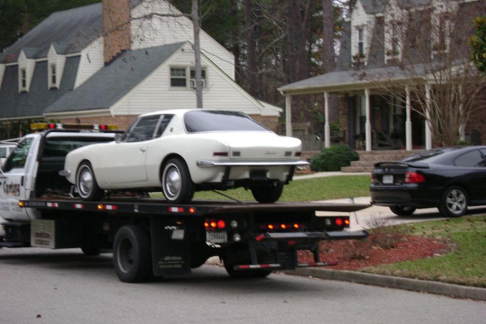 White Avanti coupe rear view on a flatbed truck with parked black car nearby