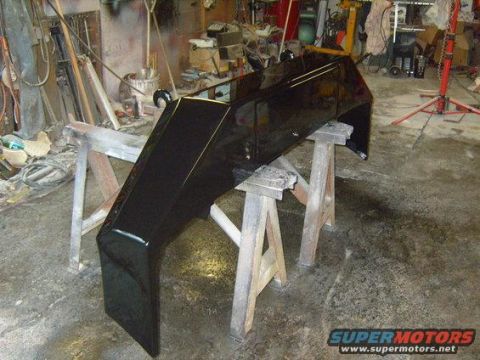 1986 Ford Bronco - A glossy black custom bumper or body kit piece is displayed on sawhorses in a workshop. The part has sharp angles and a reflective surface,…