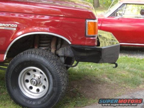 1986 Ford Bronco - The front corner of a red Ford Bronco is shown, featuring a large aftermarket black bumper and a knobby off-road tire. A red classic car is…