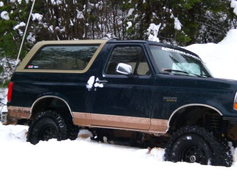1995 Ford Bronco - A dark blue Ford Bronco is stuck in deep snow, with its tires partially buried. The vehicle has a tan trim line along the lower body and a…