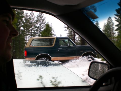 1995 Ford Bronco - A dark green Ford Bronco with a tan top drives through a snowy, watery road, kicking up spray. The photo is taken from inside another vehic…
