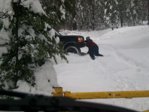 1995 Ford Bronco - A person pushes a dark-colored SUV stuck in the snow on a wooded road.