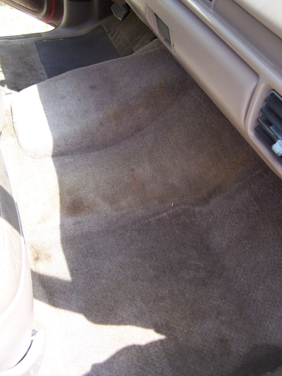 Interior of a vehicle showing a stained tan carpet floor and a black rubber mat.