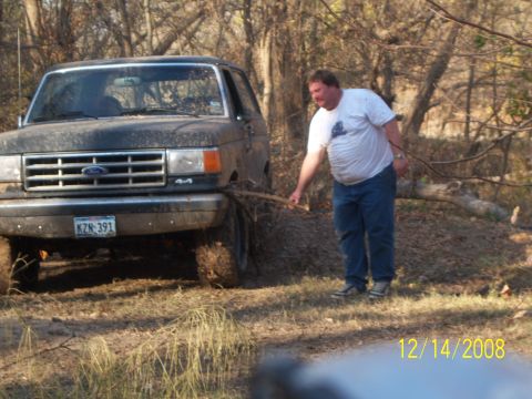 1996 Ford Bronco - A man in a white t-shirt and blue jeans is standing next to a muddy, dark-colored Ford truck that is stuck in the woods. The truck has a li…