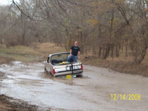 1996 Ford Bronco - A man stands in the bed of a light-colored pickup truck stuck in a muddy, wooded area. The truck is partially submerged in water and mud, w…
