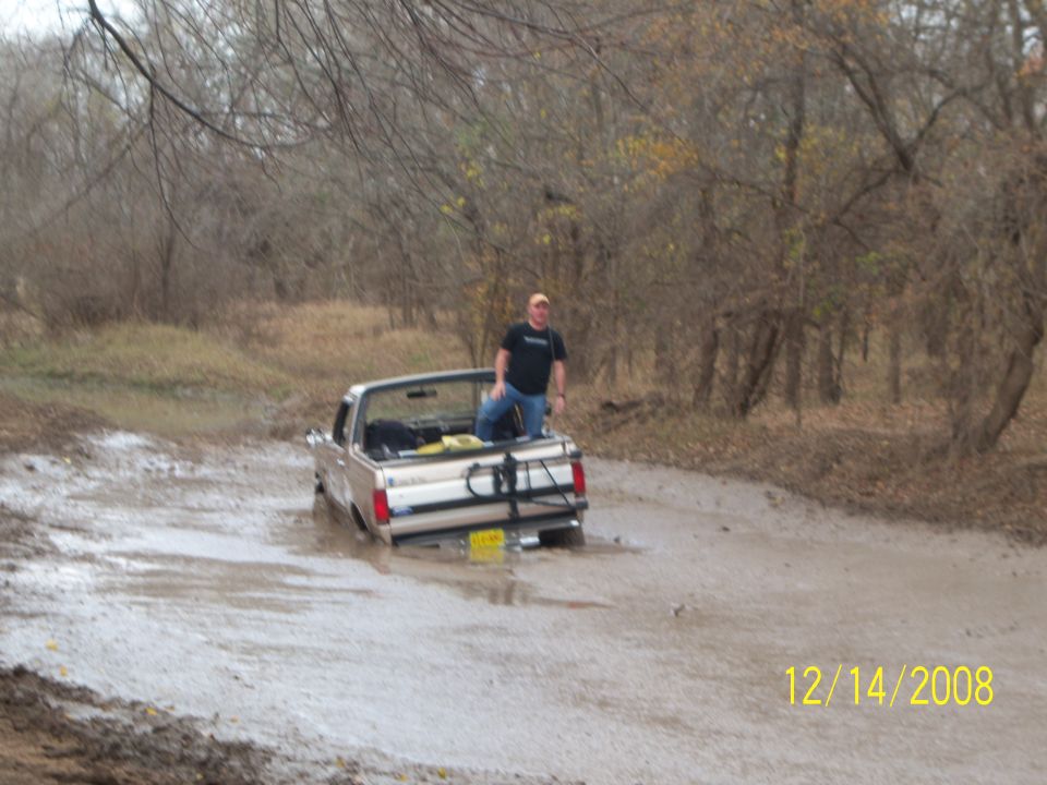 Man standing in a pickup truck stuck in mud and water in a wooded area.