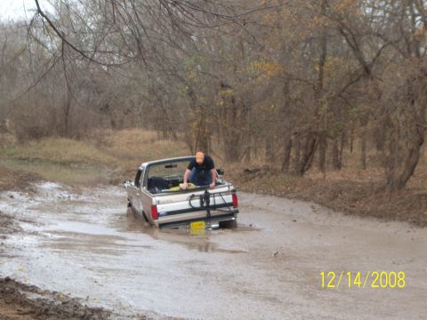 1996 Ford Bronco - A man in an orange hat is working on the bed of a light-colored pickup truck stuck in a muddy, wooded area. The truck's rear end is submerg…