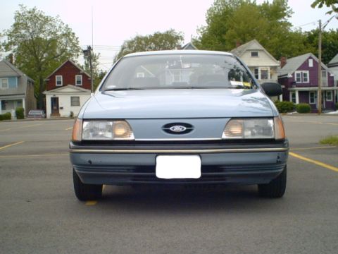 1986 Ford Taurus - Front view of a blue 1986 Ford Taurus sedan parked in a lot with houses visible behind it.