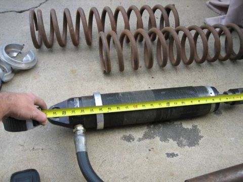 1984 Ford Bronco - A hand holds a yellow measuring tape against a black shock absorber assembly. Two large, rusty coil springs are visible in the background.