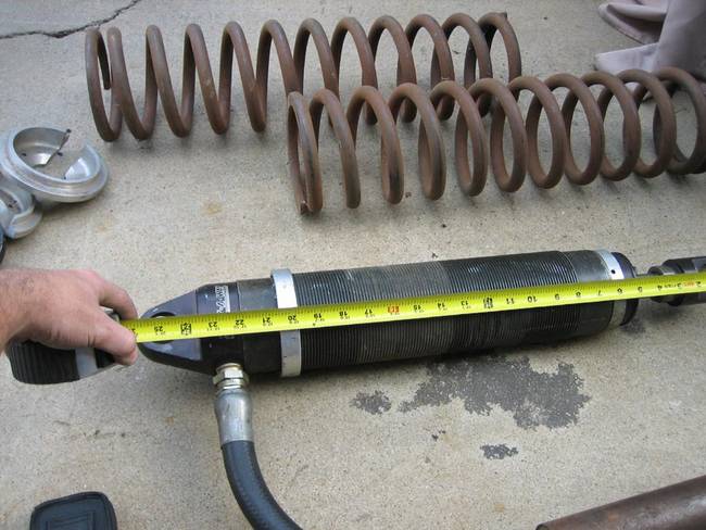 Hand measuring a black shock absorber with a yellow tape measure, rusty coil springs in background.