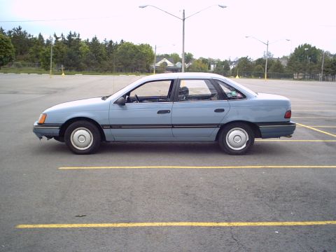 1986 Ford Taurus - Side profile of a light blue 1986 Ford Taurus sedan parked in an empty lot.