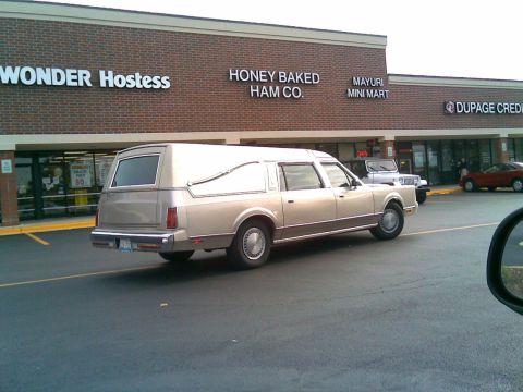 1972 Ford Ranchero - A tan hearse is parked in front of a strip mall with businesses including Wonder Hostess and Honey Baked Ham Co. The hearse is viewed from…
