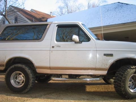 1996 Ford Bronco - Driver-side profile of a lifted white and tan two-tone Ford Bronco with the hardtop on, mud-splattered lower body, chrome nerf-bar side ste…