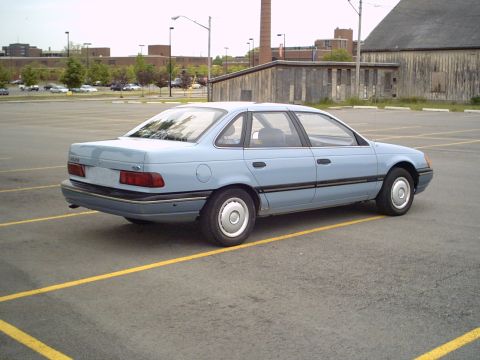 1986 Ford Taurus - A light blue 1986 Ford Taurus sedan is parked in a lot, showing its rear profile.