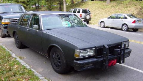 1991 Ford Crown Victoria - A matte black sedan with a push bar is parked on the side of a road next to other vehicles.