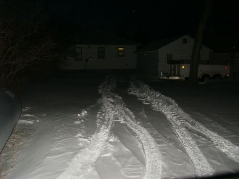 1990 Ford Bronco - Tire tracks mark a path through the snow towards houses on a dark night.