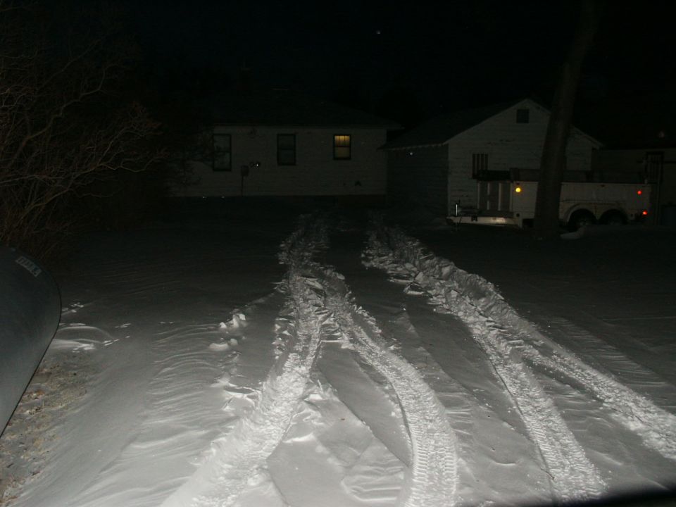 Tire tracks in snow leading towards houses at night, with a trailer visible in the background