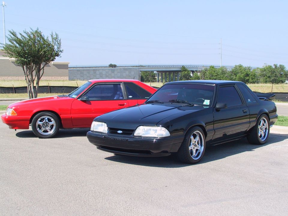 Black Fox-body Mustang coupe with chrome wheels, red Mustang and overpass behind