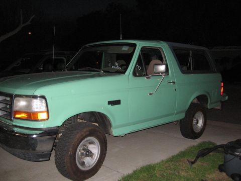 1996 Ford Bronco - A light green 1996 Ford Bronco is parked on a driveway at night. The vehicle is viewed from the side, showing the front fender, door, and r…