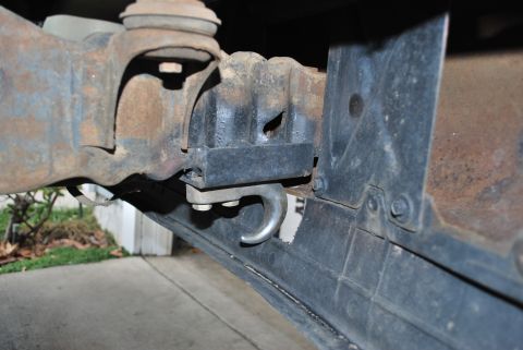 1996 Ford Bronco - A close-up view of the rusty metal frame and a black bracket with a silver tow hook attached to the underside of a vehicle.
