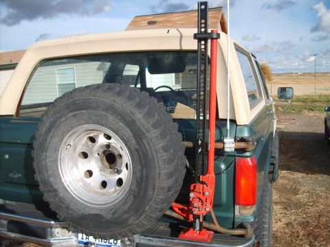 1989 Ford Bronco - The rear passenger side of a dark green 1989 Ford Bronco is shown, featuring a spare tire mounted on the back and a red Hi-Lift jack attach…