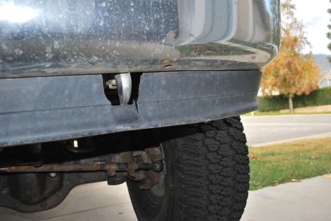 1996 Ford Bronco - A close-up view of the lower front bumper area of a vehicle, showing a torn black plastic trim piece and a chrome tow hook. The tire and pa…