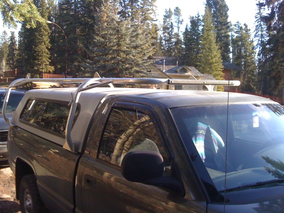Dark pickup with canopy and roof rack holding lumber, parked near pine trees.