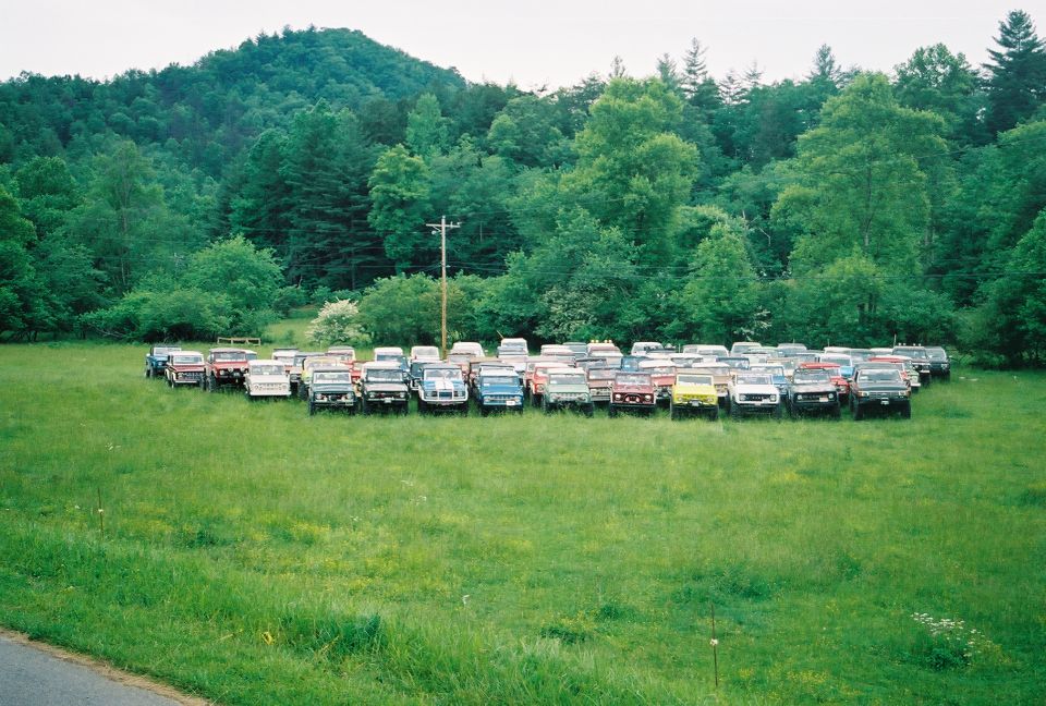 Long row of older 4x4 trucks and Broncos parked across a grassy field below wooded hills