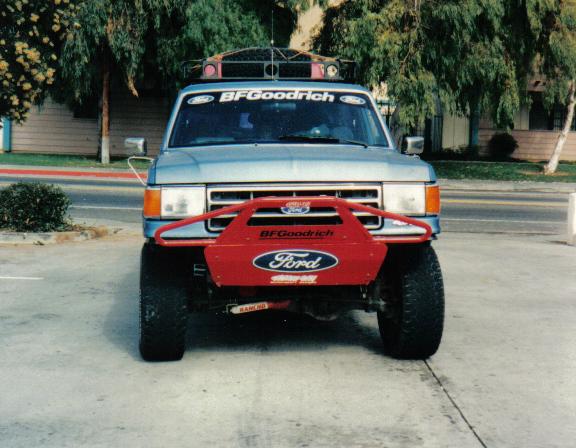 Front view of lifted blue Ford Bronco with red prerunner bumper, roof rack lights, and BFGoodrich banner