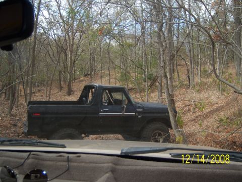 1996 Ford Bronco - A black 1990s Ford Bronco is parked on a dirt path in a wooded area. The vehicle is viewed from the interior looking out the windshield.