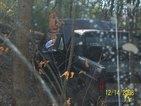 1996 Ford Bronco - A dark colored pickup truck is parked at an angle in a wooded area, with branches and leaves obscuring parts of the vehicle. The date 12/14…