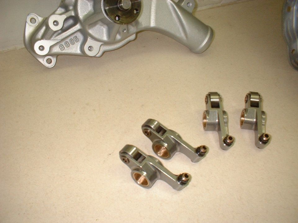 Four roller rocker arms with bronze bushings on a bench next to an aluminum water pump stamped 8805