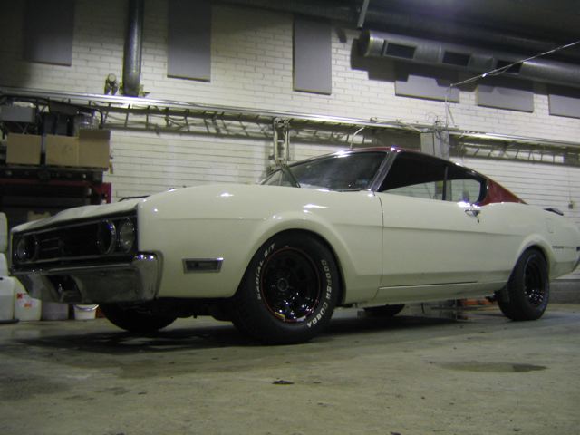 White 1960s Mercury Cyclone fastback in a shop, front bumper removed, black steel wheels