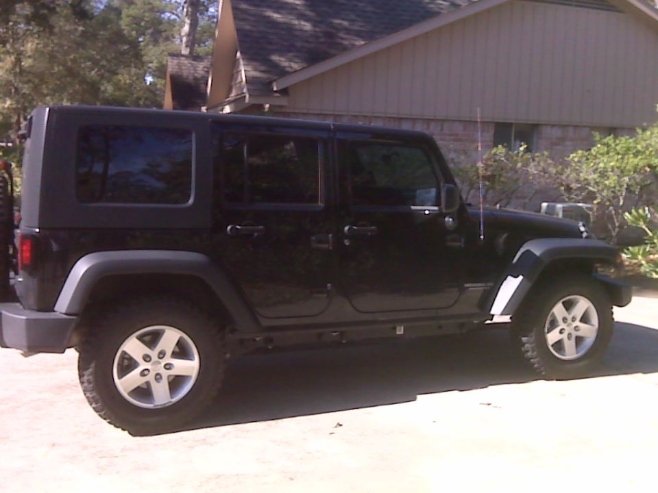 Black Jeep Wrangler, four-door model, parked outside a house. Side view shows the wheels and body