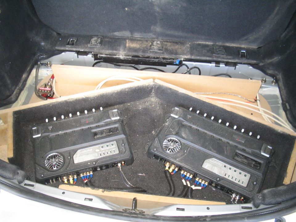 Two amplifiers on a carpeted angled rack with RCA and power wiring in a car hatch