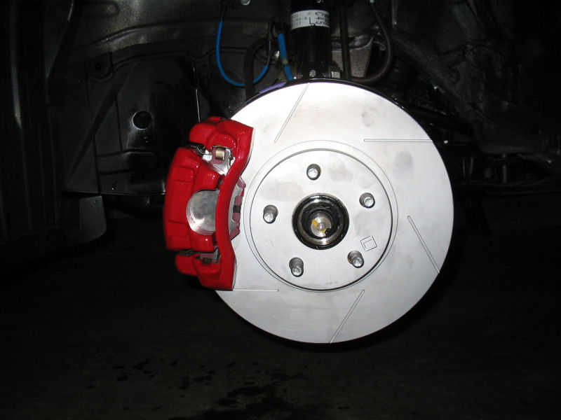 Red brake caliper over a silver vented rotor with slotted grooves, showing wheel studs and hub