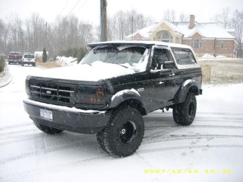 1995 Ford Bronco - A black Ford Bronco sits parked on a snow-covered road in front of a large house.