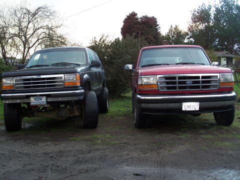 1977 Ford F-150 - A black SUV and a red pickup truck are parked side-by-side on a dirt lot with trees in the background.