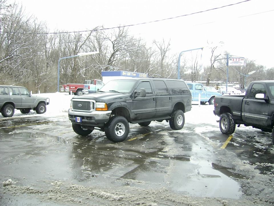 Dark Ford Excursion SUV parked in a lot with snow and puddles. Another SUV and a pickup truck are also visible