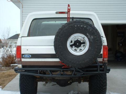 1995 Ford Bronco - The rear of a white Ford Bronco shows a spare tire, custom bumper, and a Hi-Lift jack mounted on the tailgate.