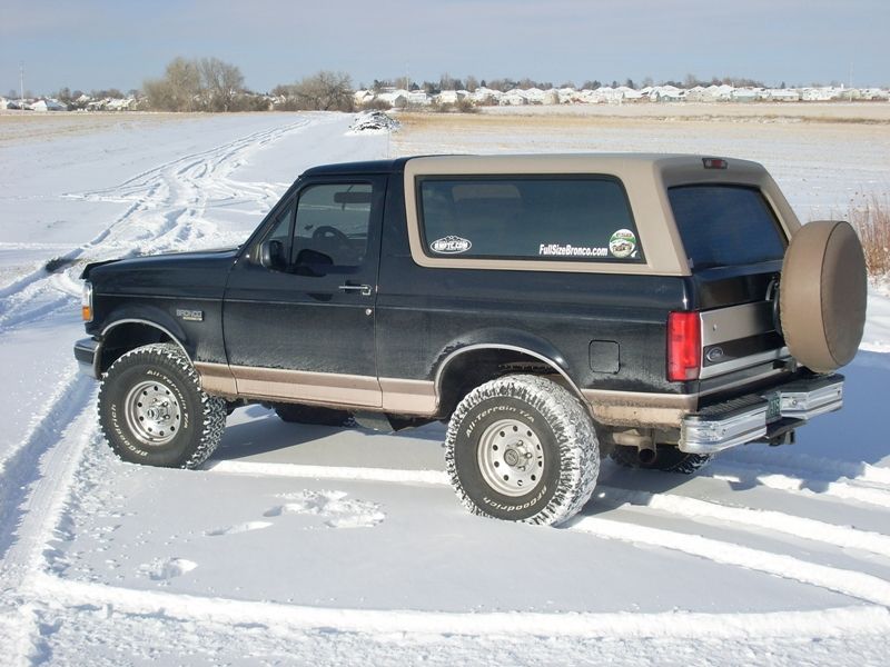 Black and tan Ford Bronco with a spare tire on the back, driving in snow