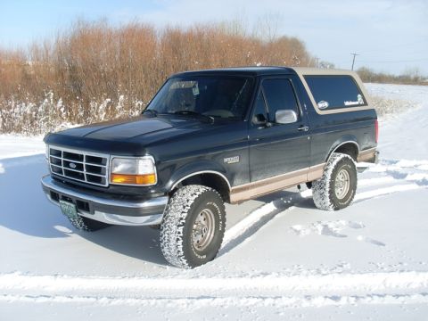 1996 Ford Bronco - Black Ford Bronco with tan lower two-tone and tan hardtop, photographed from the front three-quarter on a snow-covered field under blue sky…
