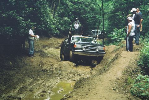 1991 Ford Bronco - Several people watch as a black Ford F-Series truck drives through a muddy off-road path.