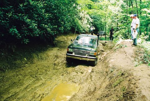 1991 Ford Bronco - A black SUV navigates a muddy trail in a wooded area with onlookers.